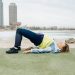A woman in activewear exercises near the beach with skyscrapers in the background, embodying a healthy lifestyle.